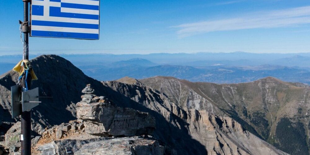 Auf dem Mytikas Gipfel mit griechischer Flagge im Vordergrund und dem Olymp Gebirge im Hintergrund